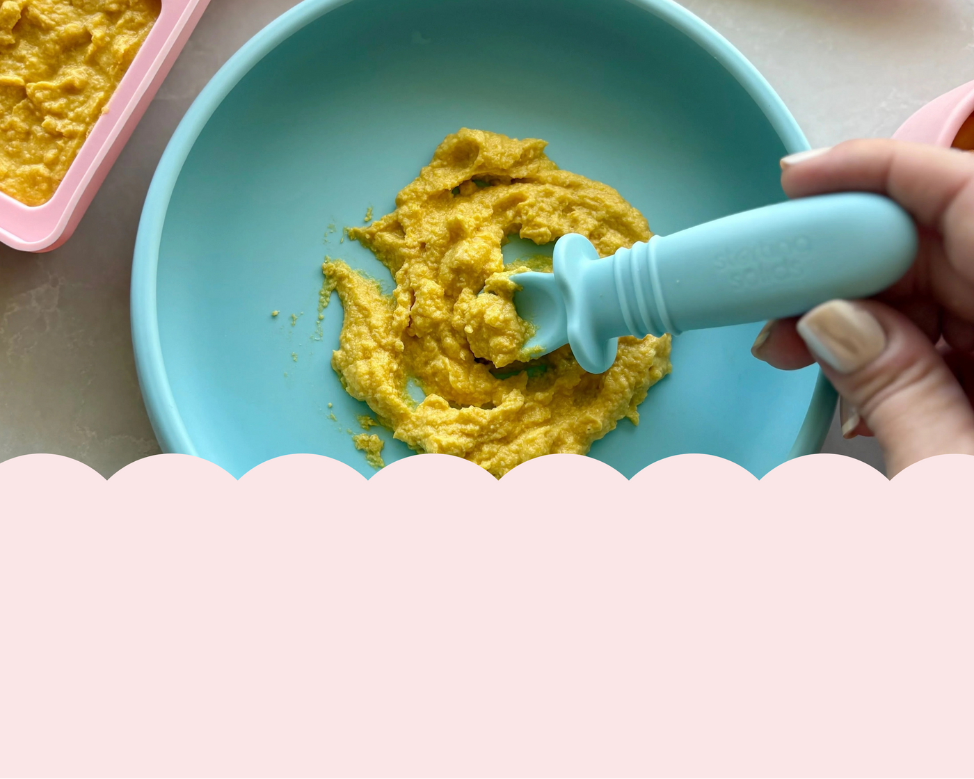 Yellow baby food in a blue bowl with a pink baby food tray and carrot in the background.
