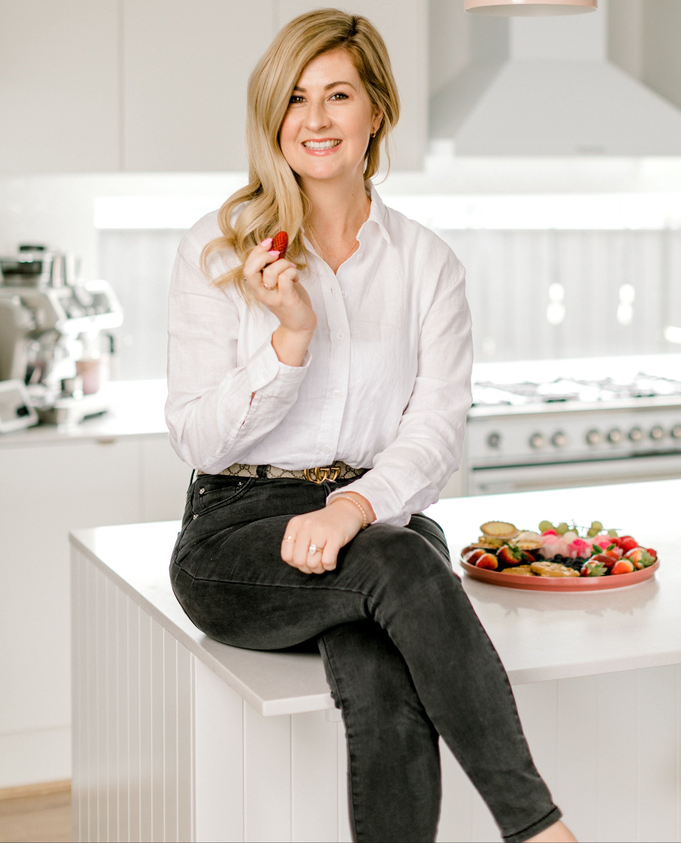 Woman sitting on a kitchen counter holding a piece of fruit, with a modern kitchen in the background.