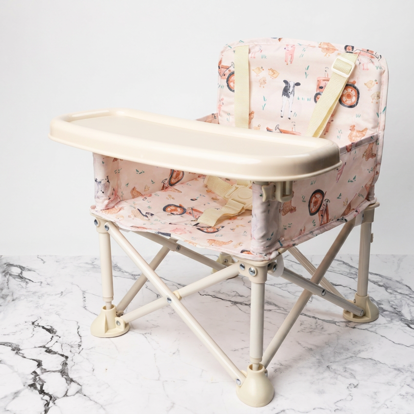 A portable baby high chair with a cream-colored tray and frame, upholstered in a pink and beige cartoon animal print fabric, shown on a white marble surface against a white background.