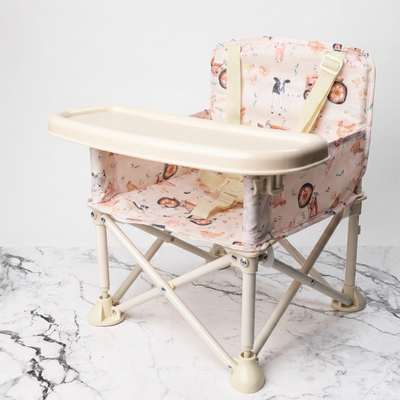 A portable baby high chair with a cream-colored tray and frame, upholstered in a pink and beige cartoon animal print fabric, shown on a white marble surface against a white background.