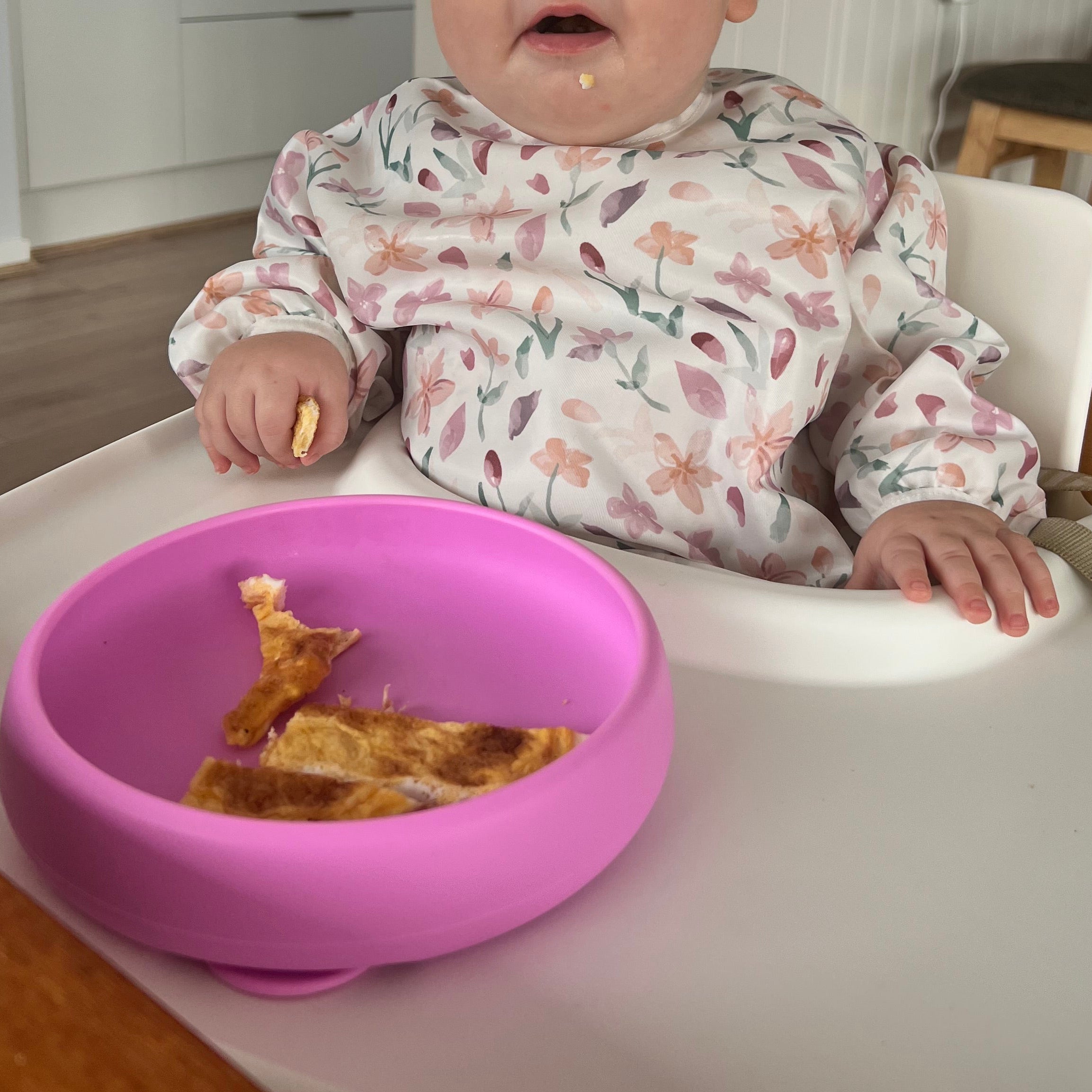 Baby in a high chair with a pink tray holding food, in a home setting.