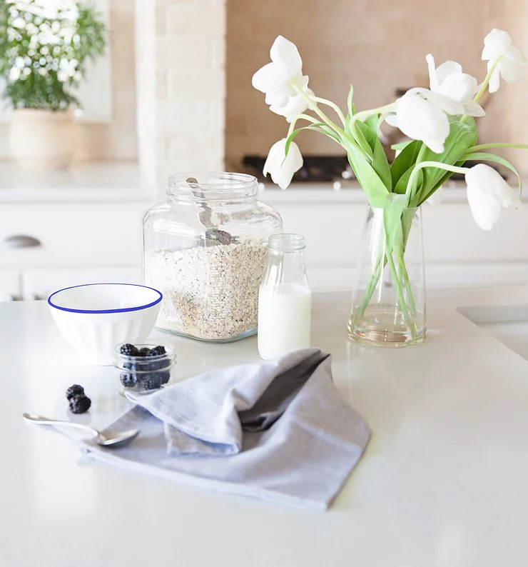 A jar of oats, a bowl, milk, berries, spoon and tea towel on a kitchen bench with a vase of white tulips - Starting Solids Australia recipe for porridge.