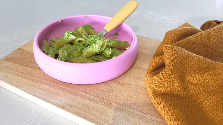 Iron rich pesto with penne pasta in pink silicone suction plate with yellow baby fork on a wooden board - Starting Solids Australia recipe.