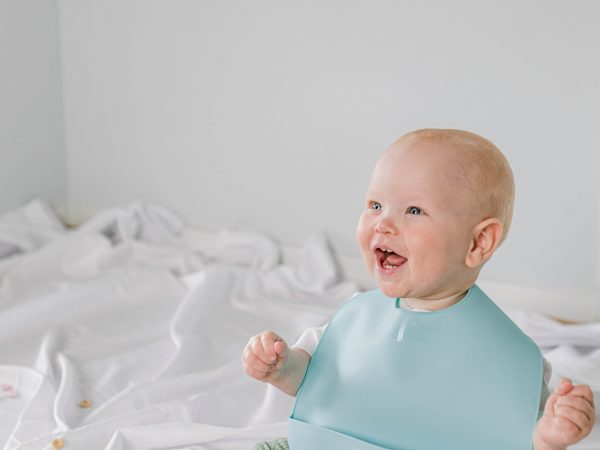 Happy baby sitting on white sheet wearing Starting Solids Australia's silicone catch-all 'Silly Bib'.