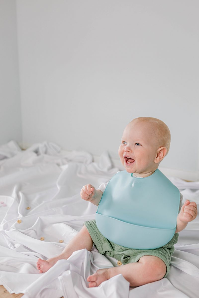 Happy baby sitting on white sheet wearing Starting Solids Australia's silicone catch-all 'Silly Bib'.