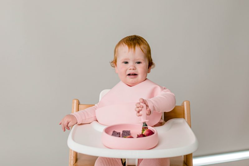 Baby girl sitting in Tripp Trapp highchair wearing Starting Solids Australia silicone Silly Bib and eating strawberries with Toddler Silvies out of a Suckie Scoop Plate.