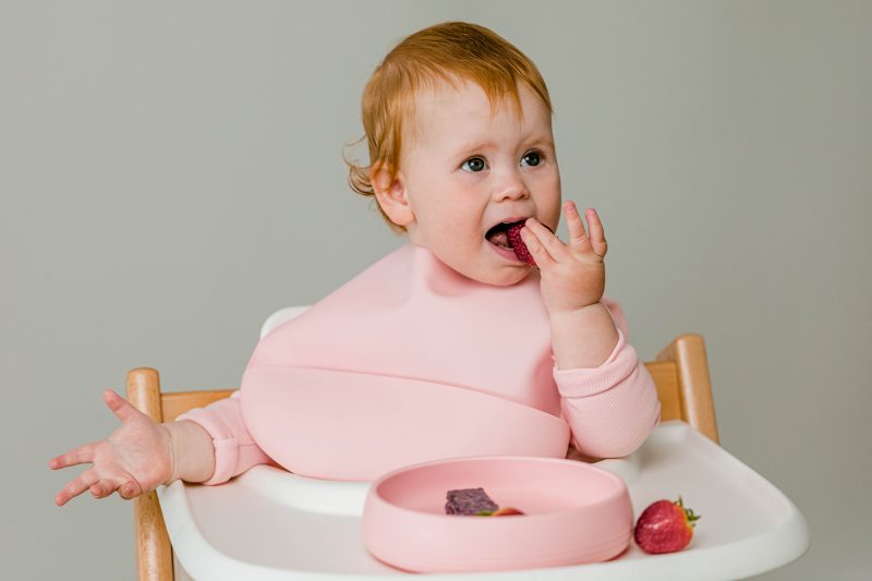 Baby girl wearing Starting Solids Australia silicone Silly Bib in Trapp Trapp high chair, and eating food from a Suckie Scoop Plate.