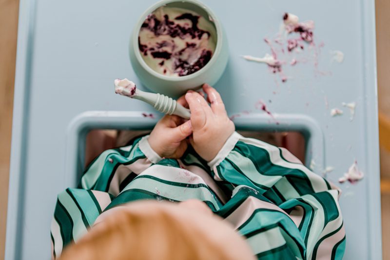 Birdseye view of baby in highchair eating berries and yoghurt from a Suckie Scoop Bowl with a Sticky Spoon, wearing a Smockie Bib - all by Starting Solids Australia.