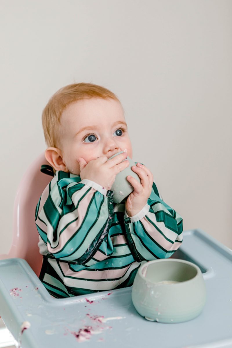 Baby boy sitting in a highchair wearing Starting Solids Australia's Smockie Bib in Aqua Lines print, eating yoghurt from a Suckie Scoop Bowl and Bubbie Cup.