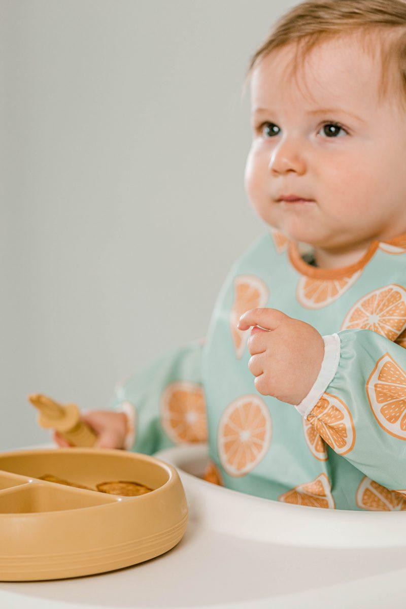Baby wearing Starting Solids Australia's Smockie Bib in Oranges print whilst eating solid food with a silicone Selfie Spoon from a Suckie Scoop Divided Plate.