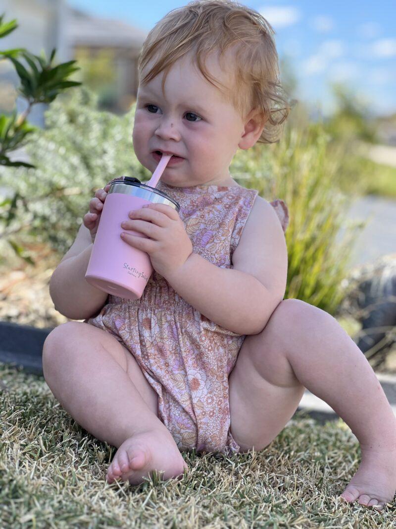 Toddler girl sitting on grass drinking from a pink stainless steel insulated 'Big Kiddie Cup' with straw for kids by Starting Solids Australia.