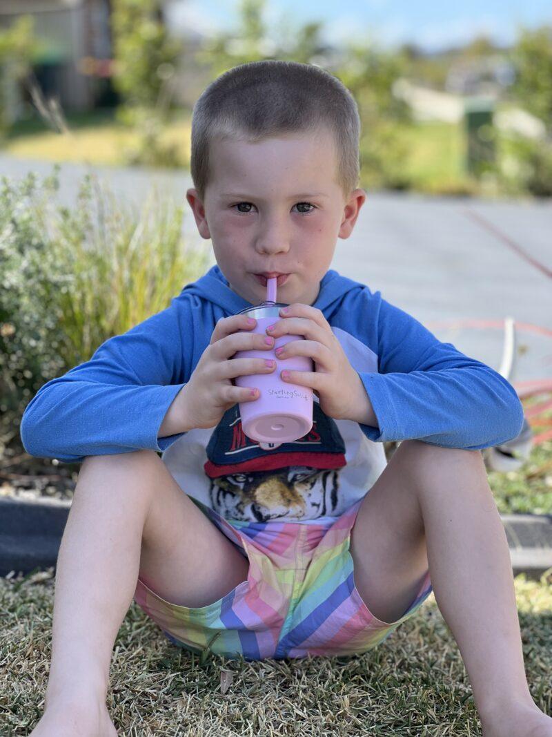 Young boy sitting on grass drinking from a pink stainless steel insulated 'Big Kiddie Cup' with straw by Starting Solids Australia.