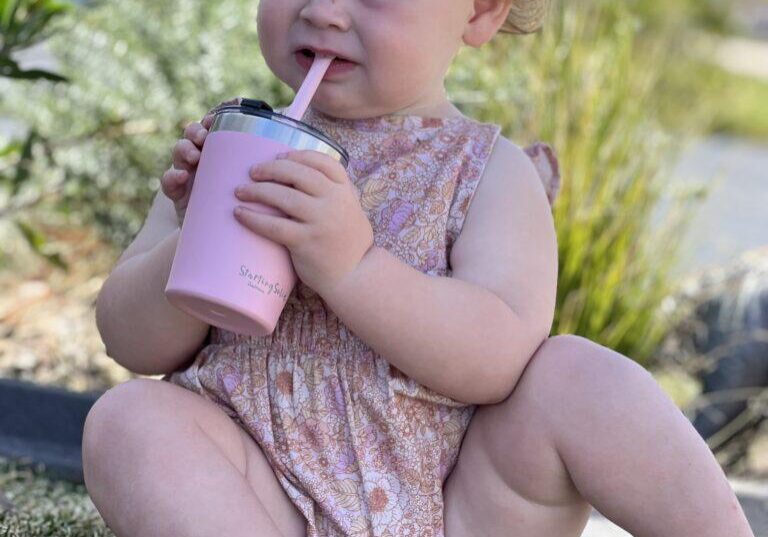 Toddler girl sitting on grass drinking from a pink stainless steel insulated 'Big Kiddie Cup' with straw for kids by Starting Solids Australia.
